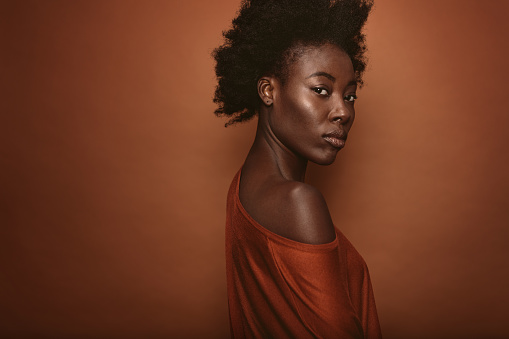 Portrait of young woman with afro hairstyle standing against brown background. African female model looking at camera.