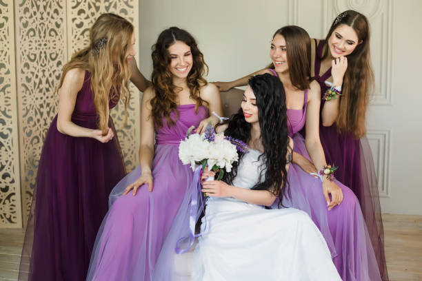 Felling excited about wedding. Attractive young bride holding a wedding bouquet and smiling while talking to her charming bridesmaids in purple dresses