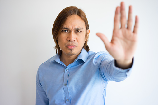 Portrait of serious young Asian man with hand raised in stop gesture. Freelancer warning colleagues of making typical mistakes. Stop and warning concept