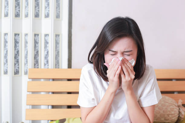 Asian woman blowing her nose while sitting on bed. illness woman sneezing in a tissue. stock photo