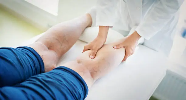 Closeup side view of unrecognizable female doctor massaging legs and calves of a senior female patient with visible varicose veins.