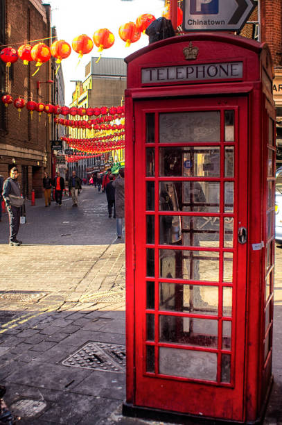 Chinese lanterns hung over Wardour Street in China Town, London during Chinese New Year. They have Chinese text on them and are backlit by late afternoon sunlight.  In the foreground is a red London telephone box.