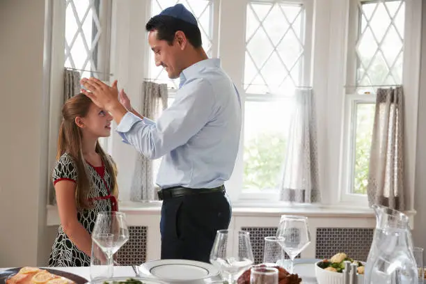 Photo of Jewish father blesses daughter by table set for Shabbat meal