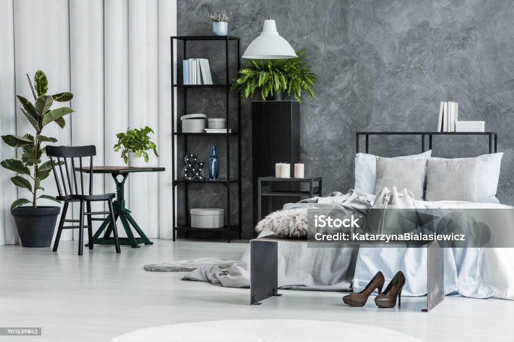 Black chair in bright bedroom Black chair at table with plant in bright bedroom with bench and high heels in front of bed against concrete wall Apartment Stock Photo