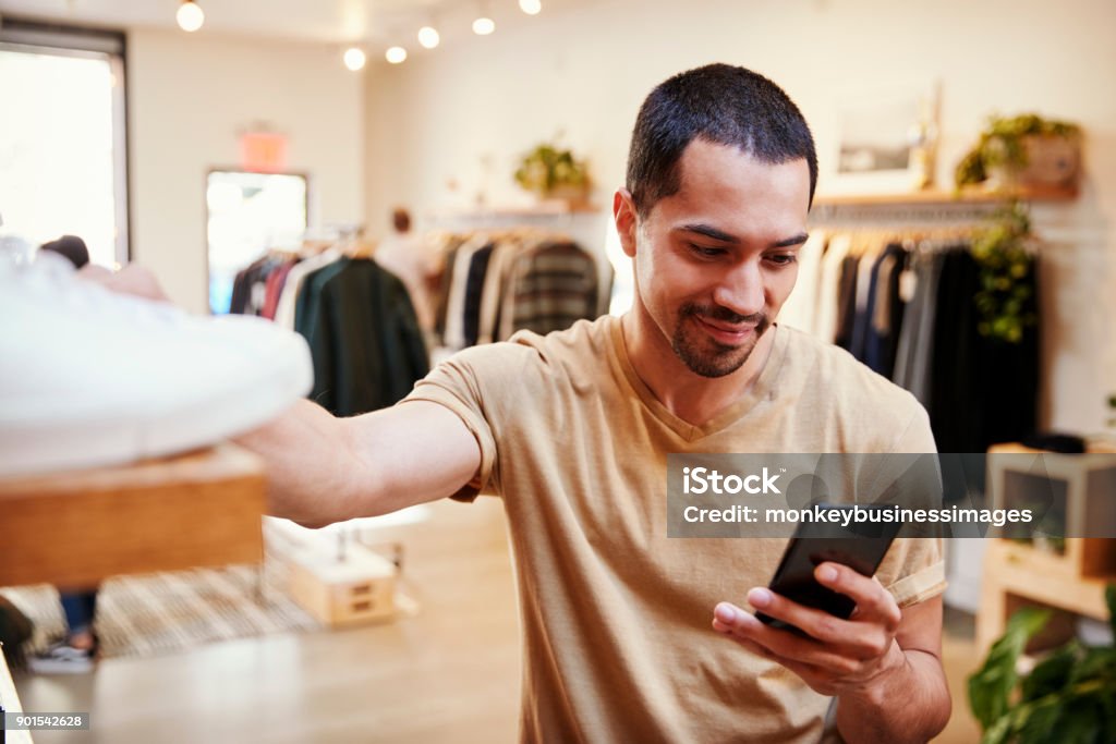 Sonriente hombre hispánico con smartphone en una tienda de ropa - Foto de stock de Venta al por menor libre de derechos