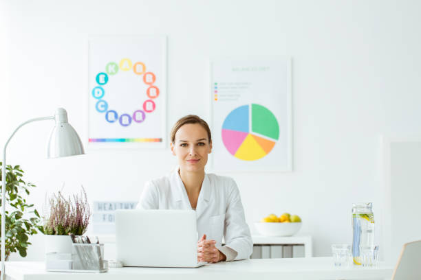 Expert on diet and nutrition Smiling expert on diet and nutrition sitting at desk with laptop and lamp in white office with posters general military rank stock pictures, royalty-free photos & images