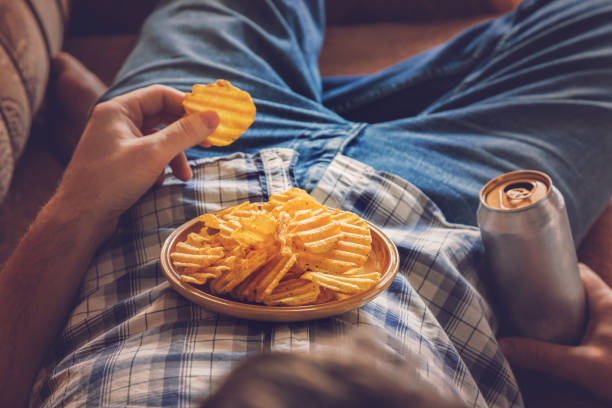 After work a guy wearing shirt and jeans lying on sofa, drinking a cold beer, eating crisps and watching sport tv channel. Man's resting time at home concept. After work a guy wearing shirt and jeans lying on sofa, drinking a cold beer, eating crisps and watching sport tv channel. Man's resting time at home concept. unhealthy eating stock pictures, royalty-free photos & images