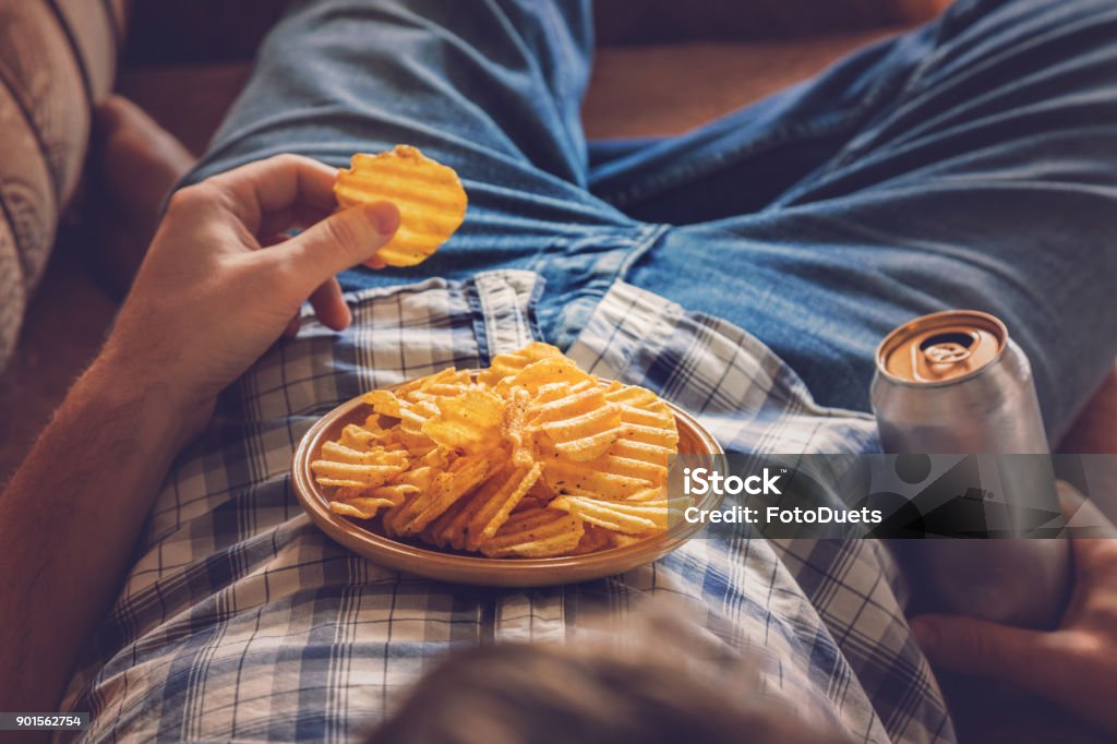 下班後, 一個男人穿著襯衫和牛仔褲躺在沙發上, 喝著冰啤酒, 吃薯片和看體育電視頻道。人的休息時間在家裡的概念。 - 免版稅不健康飲食圖庫照片
