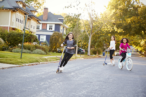 Three pre-teen girls playing in street on scooters and bike