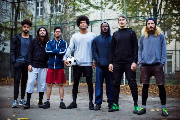 Full length of friends standing against fence. Confident soccer players with ball in court. They are in sports clothing at city.