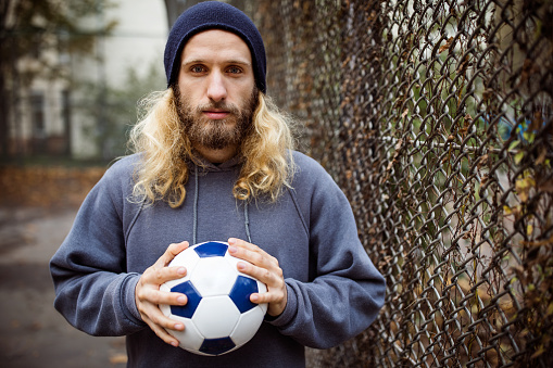 Portrait of young man holding soccer ball. Serious player is standing by chainlink fence. He is having long blond hair.