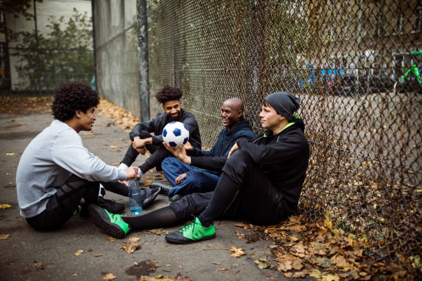 Soccer players talking while sitting against fence Soccer players talking while sitting on street. Male is holding ball while resting with team against fence. They are in sportswear during autumn. street friends stock pictures, royalty-free photos & images