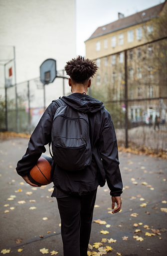 Rear view of player holding basketball. Young male is carrying backpack. He is walking on street during autumn.
