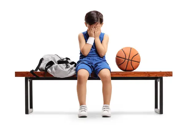 Sad little basketball player sitting on a bench isolated on white background