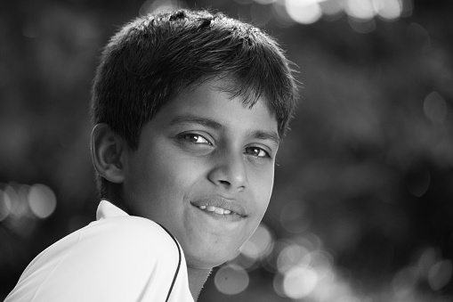 Close-up of 7 year old boy with short black hair wearing casual t-shirt and standing in sunny courtyard of family home smiling at camera.