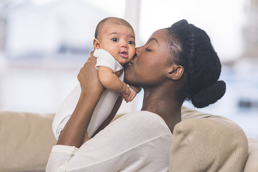 A beautiful young African American mother gently holds her infant daughter up in the air with both hands and kisses her cheek. The baby's eyes are wide open and she looks happy. They are sitting on a couch in their living room.