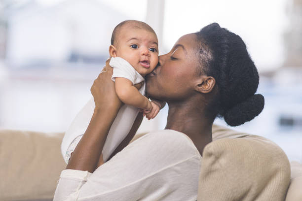 Beautiful Ethnic Mom Holds Her Infant Girl A beautiful young African American mother gently holds her infant daughter up in the air with both hands and kisses her cheek. The baby's eyes are wide open and she looks happy. They are sitting on a couch in their living room. baby goods stock pictures, royalty-free photos & images