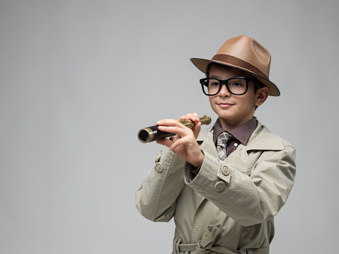 Portrait of curious little boy wearing a beige trench coat , horn rimmed glasses and a brown fedora hat looking through hand held telescope. The background is gray. Shot in studio with a medium format camera.