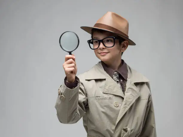 Portrait of curious little boy wearing a beige trench coat , horn rimmed glasses and a brown fedora hat looking through  magnifying glass. The background is gray. Shot in studio with a medium format camera.