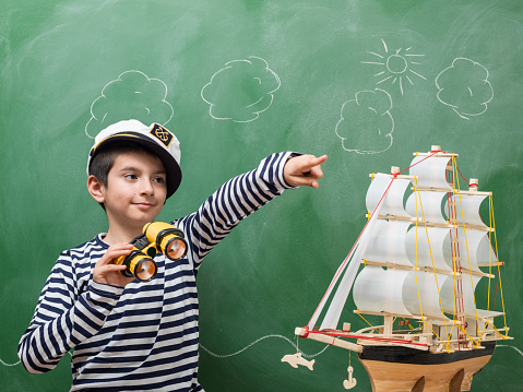 Portrait of little boy wearing a striped sailors shirt and hat holding binoculars and pointing forward. A toy ship model is seen next to him. He is seen in front of green chalkboard. Cloud and sky sketch is seen on blackboard. Shot indoor with a medium format camera.