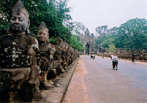 locals on motorbikes pass ancient statues line entrance gate to the ancient city of angkor cambodia 