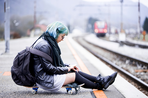 Young hipster girl in causal clothes with green hair, sitting on a skateboard by the railroad track and using her smart phone.