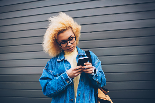 Pretty hipster girl checking smartphone notification standing on wall promotional background, thoughtful young woman checking money on banking account via phone application spending time outdoors