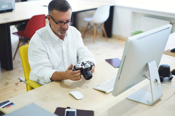 istock Mature experienced architector in eyeglasses looking photos from building office planning on vintage camera for understanding and developing future project sitting on workplace at modern computer 901746380