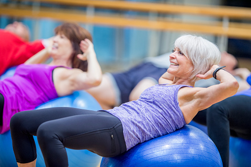 A group of adults are indoors in a fitness center. They are wearing casual athletic clothing. They are lying on exercise balls and doing core exercises.