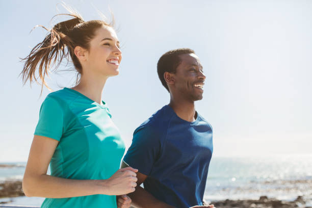 Smiling couple jogging by sea against clear sky. Man and woman are exercising on sunny day. They are in sports clothing.