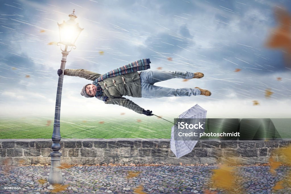 Man gets blown away by a storm Man flying away horizontally in a storm while holding on to a street lamp with one hand. The other hand is holding an upended umbrella. Wind Stock Photo