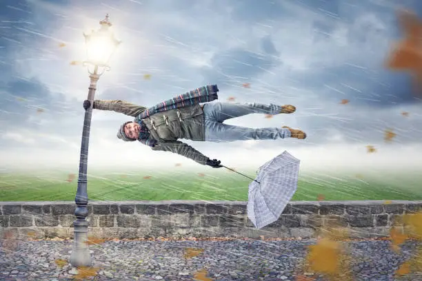 Man flying away horizontally in a storm while holding on to a street lamp with one hand. The other hand is holding an upended umbrella.