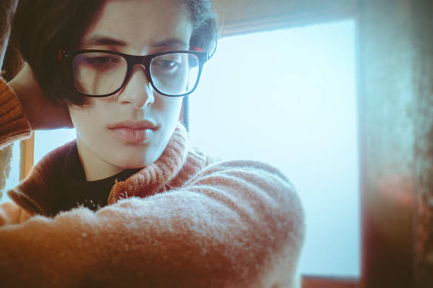 Indoor image of disturbed young woman sitting near door and thinking. She is wearing eyeglasses and looking down. One person, headshot and selective focus with copy space.