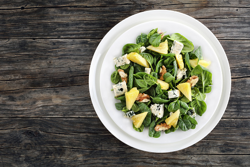 fresh Baby spinach, pineapple slices, blue cheese chunks and walnuts salad on a white plate on dark wooden table, view from above, close-up