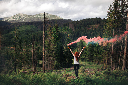 Young Caucasian woman standing in the forest in mountains  with pink and green smoke bomb in hands
