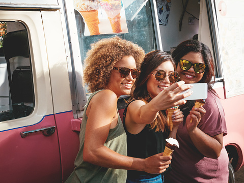 Three beautiful diverse young women eating ice cream near the ice cream truck and taking selfie