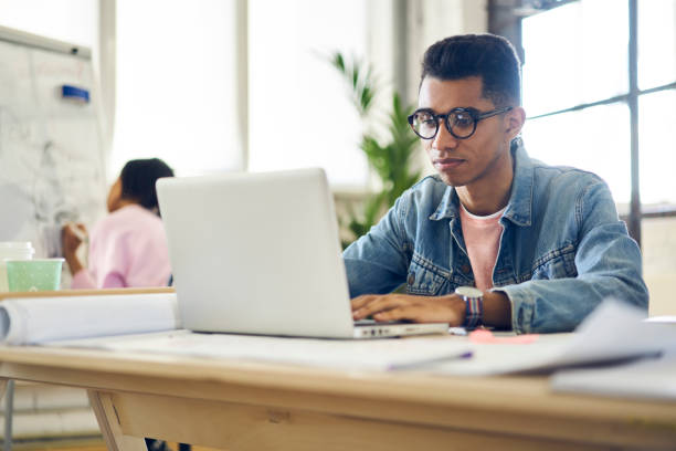 concentré d’afro américain élève de sexe masculin de celui-ci académie en programmation sur ordinateur portable en studio intérieur de loft, informations de navigation freelance qualifiés sur le site web développement de logiciels de lunettes tendance - lundi noir chinois photos et images de collection