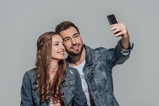 happy young couple in denim jackets taking selfie with smartphone isolated on grey