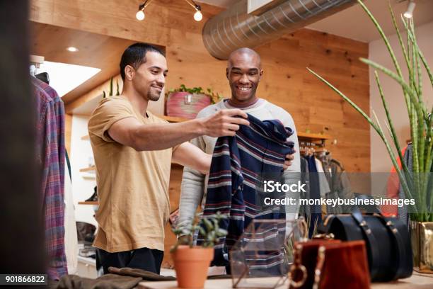 Two Young Men Holding Up Clothes To Look At In Clothes Shop Stock Photo - Download Image Now
