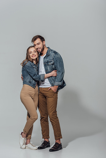 full length view of cheerful young couple hugging and smiling at camera isolated on grey