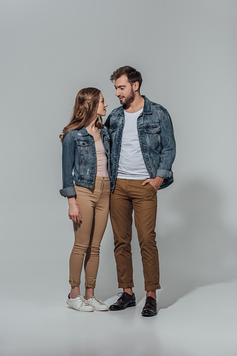 full length view of smiling young couple in denim jackets looking at each other isolated on grey