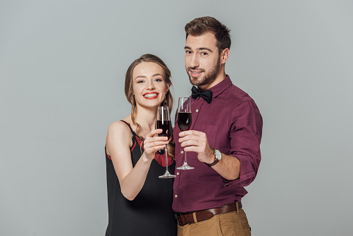 beautiful elegant young couple holding glasses of red wine and smiling at camera isolated on grey