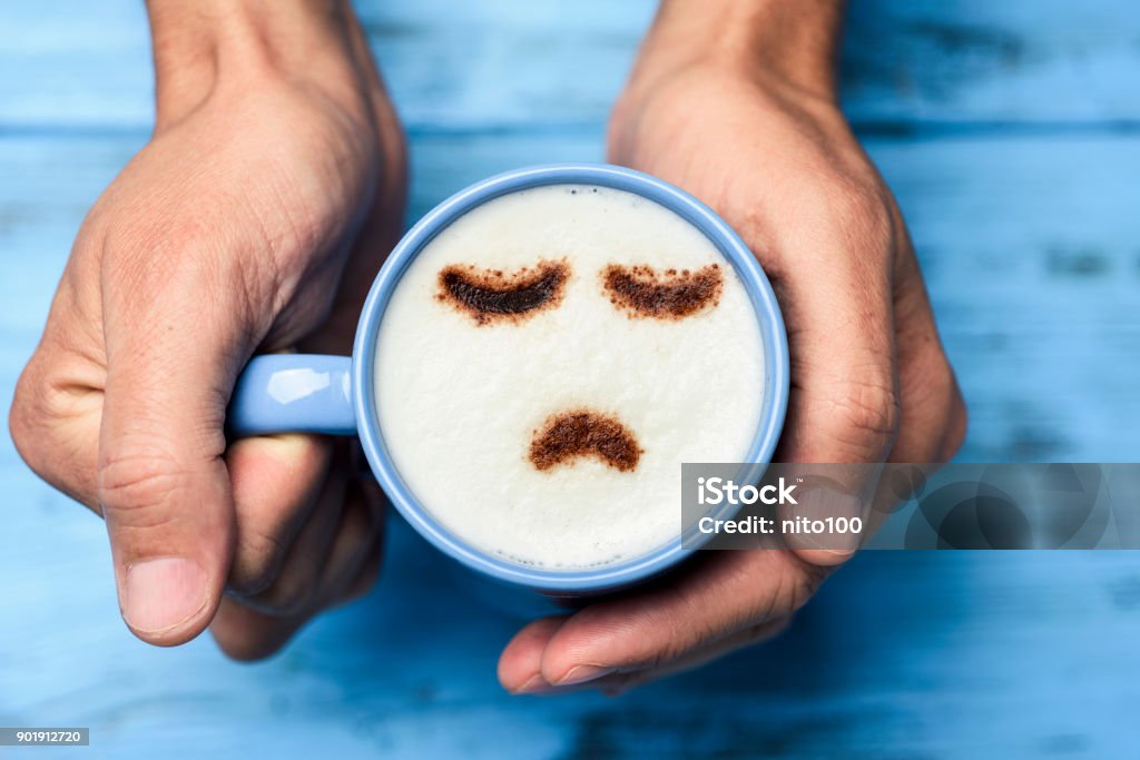 man with a cup of cappuccino with a sad face high-angle shot of a young caucasian man with a blue cup of cappuccino with a sad face drawn with cocoa powder on the milk foam, on a blue rustic table Blue Monday - Date Stock Photo