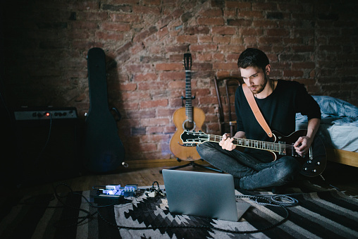 A young musician is playing a guitar in a bedroom. He is performing in front of a laptop. The room is dark but cozy.