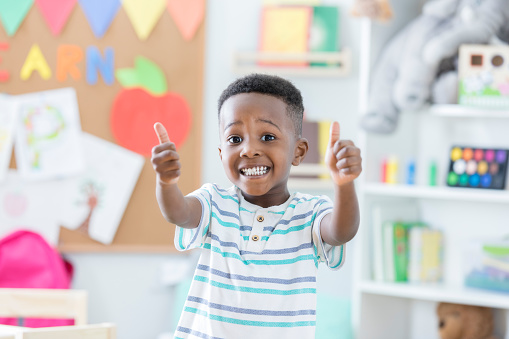 An adorable little boy smiles for the camera in his new day care center.  He holds two thumbs up to share his opinion of the place.