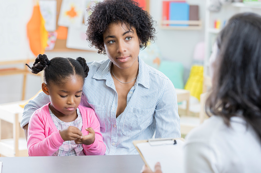 A young mother holds her preschool age daughter in her lap as she sits across a table from her daughter's teacher and listens to her testing evaluation.
