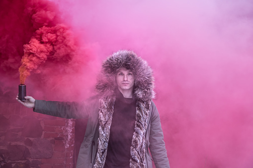 Young caucasian man in a green winter parka posing with a red smoke bomb on a sombre winter day. Tartu, Estonia.