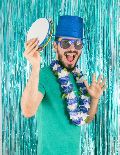 Man is playing the tambourine. Musical instrument. Bearded Brazilian is wearing green T-shirt, blue top hat and Hawaiian necklace. Carnival in Brazil, Party, Joy, New Year's Eve."n Young Man is showing a musical instrument: tambourine. Dressed up. Bearded Brazilian is wearing green T-shirt, blue top hat and white Hawaiian necklace. Bright and colorful background. Carnival in Brazil, Party, Joy, New Year's Eve, Mardi Gras. samba dancing stock pictures, royalty-free photos & images