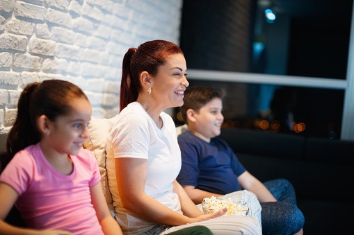 Movie night at home with divorced mother, daughter and son. Modern family watching television and eating popcorn, sitting on sofa at home. They laugh while watching comedy show on TV