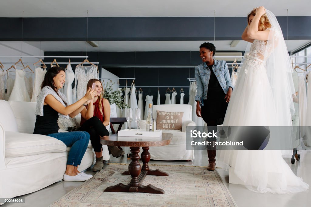 Woman trying on wedding dress in a shop with friends Woman trying on wedding dress with female friends having fun and taking photographs. Wedding Dress Stock Photo